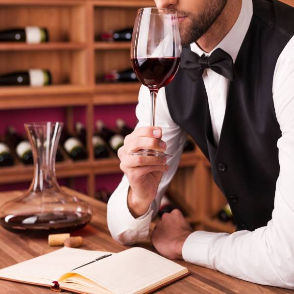 Sommelier examining wine. Cropped image of confident male sommelier examining wine while smelling it and leaning at the wooden table with wine shelf in the background