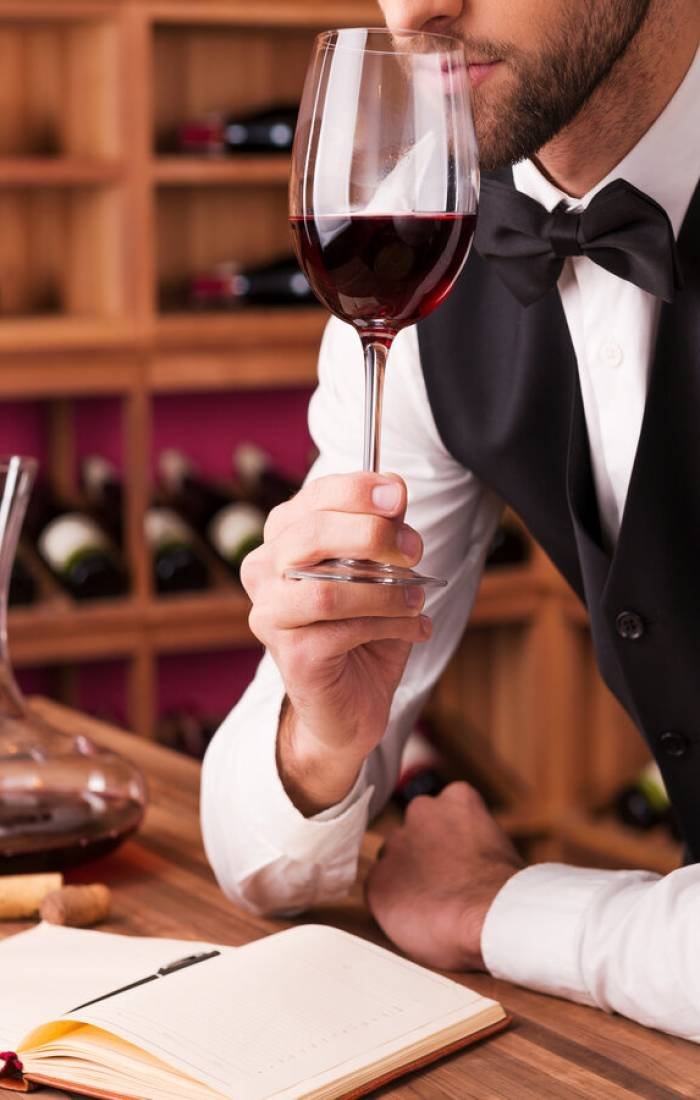 Sommelier examining wine. Cropped image of confident male sommelier examining wine while smelling it and leaning at the wooden table with wine shelf in the background